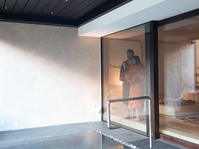 Val di Fassa in estate o inverno? Il relax non ha tempo! Coppia abbracciata all'interno di una stanza con vista su una piscina coperta