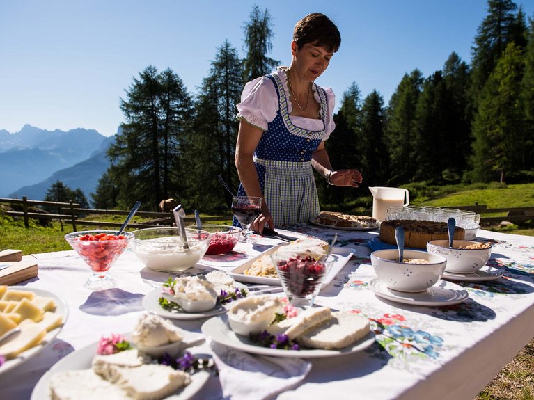 La pista ciclabile in Val di Fassa? C’è molto di più! Donna in abito tradizionale prepara cibo su tavolo all'aperto in montagna