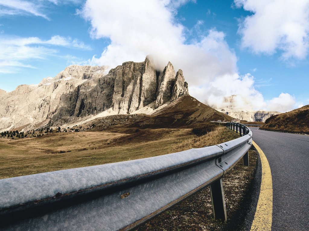 Noleggio bici a Soraga: un must in autunno all’Hotel Latemar Strada curva con guardrail vicino a montagne rocciose e cielo azzurro con nuvole
