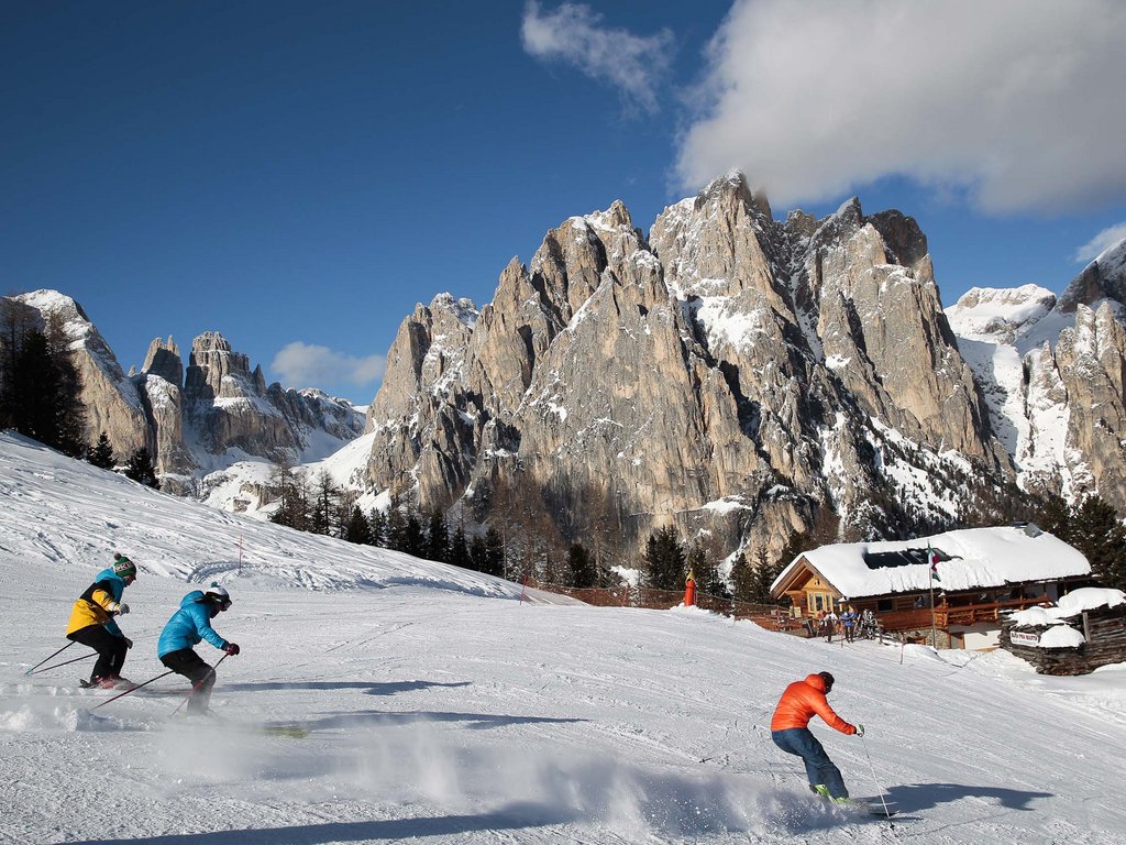 Passeggiate invernali in Val di Fassa: Hotel Latemar Sciatori che scendono una pista innevata con montagne rocciose sullo sfondo