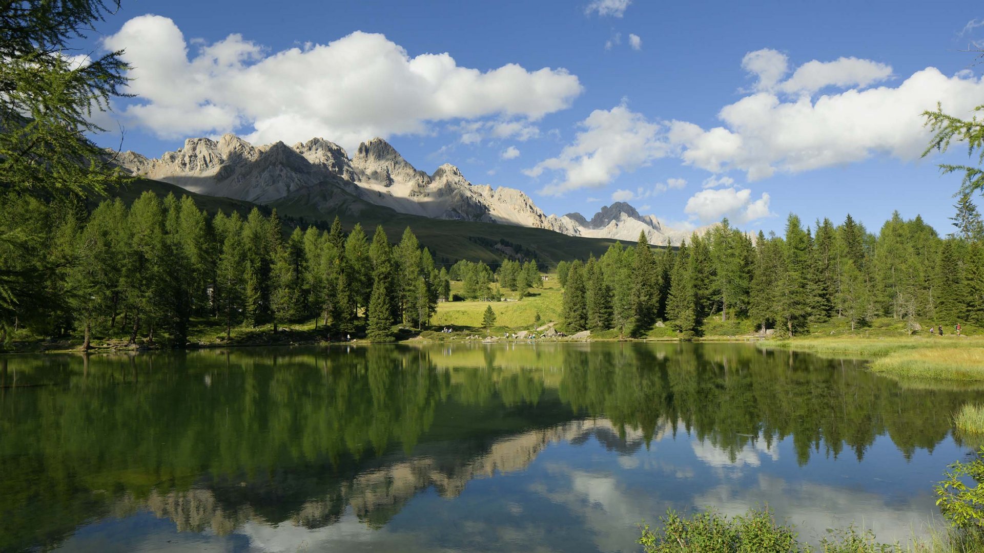 Val di Fassa: dove dormire e vivere vacanze sostenibili Lago alpino tranquillo con riflessi di alberi e montagne soleggiate