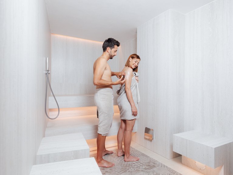 Il rituale dell’hammam al Latemar Hotel Coppia in accappatoio in una spa con pareti e pavimento in marmo chiaro.