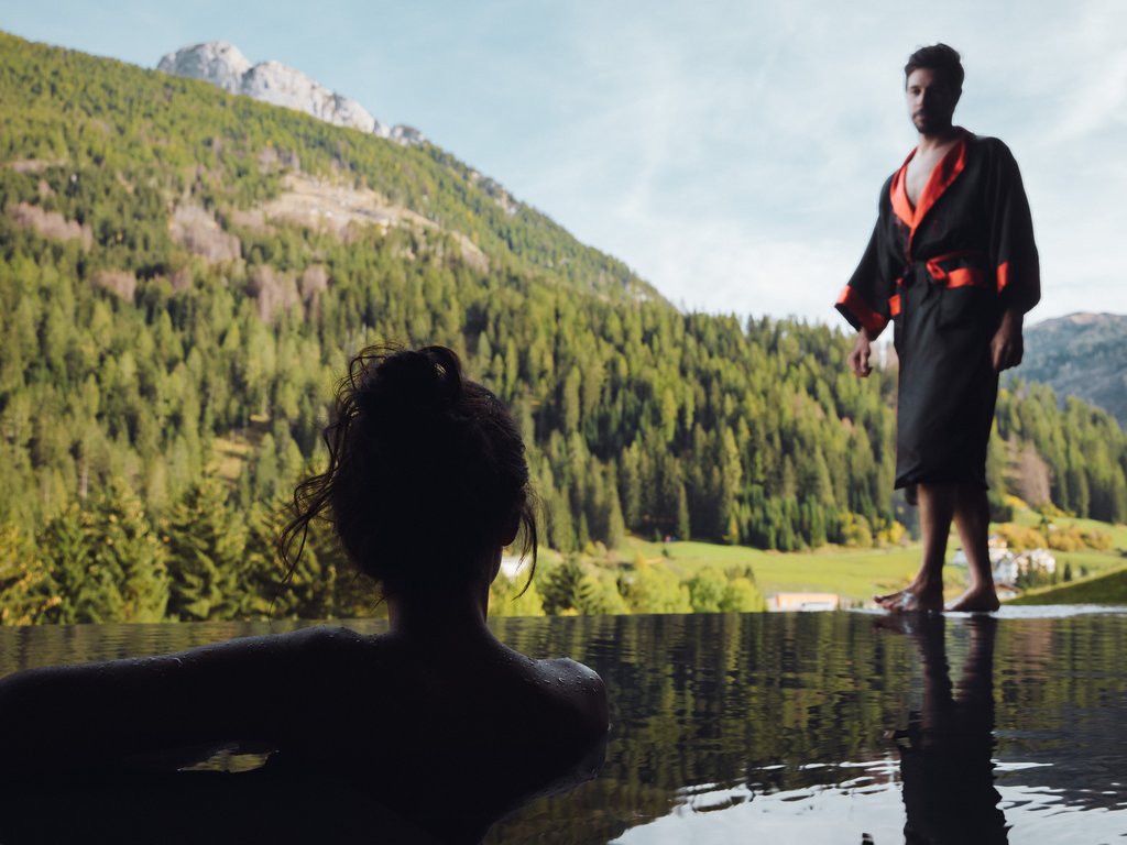 Sulla pista ciclabile, da Moena alla vostra oasi romantica Donna in piscina e uomo in accappatoio con vista sulle montagne