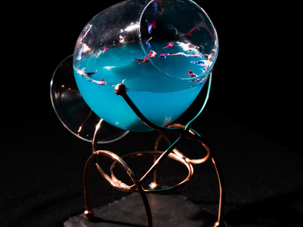 Val di Fassa e ciclabile: da ammirare dal cocktail & tea bar Bicchiere inclinato con liquido blu e petali colorati su supporto metallico