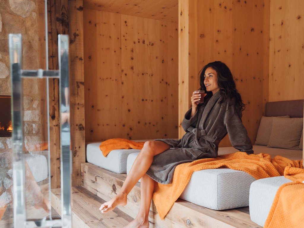Al Latemar, dopo le passeggiate: relax nella Spa Oniro Donna in accappatoio grigio che beve bevanda, seduta in ambiente in legno tipico di spa