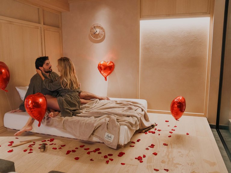 Val di Fassa in estate o inverno? Il relax non ha tempo! Coppia romantica su letto con palloncini a forma di cuore e petali rossi