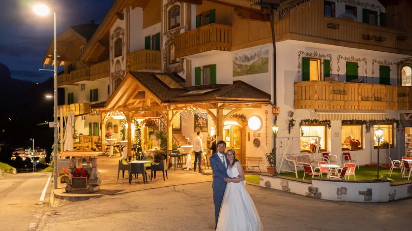 La pista ciclabile in Val di Fassa? C’è molto di più! Coppia di sposi davanti a un ristorante illuminato di sera.
