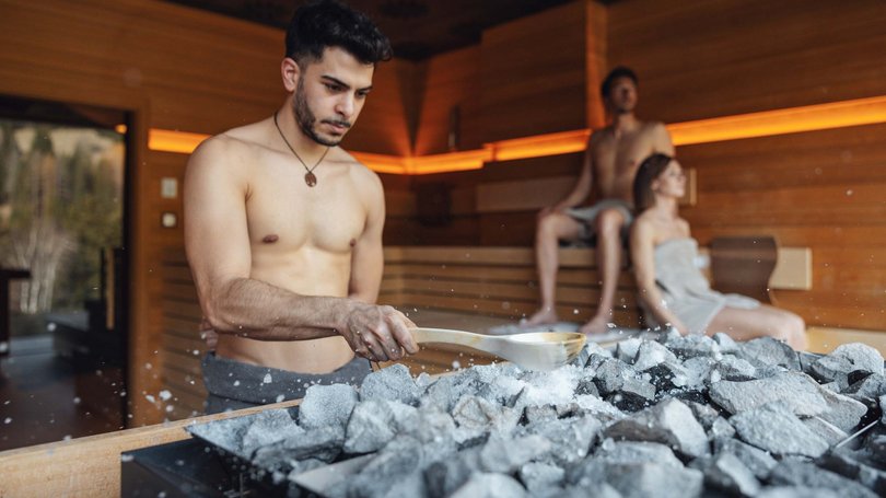 Escursioni da Moena e molto altro: il nostro programma Uomo versa acqua sulle pietre calde in sauna con due persone sedute sullo sfondo