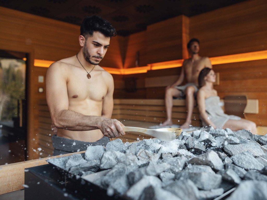 Al Latemar, dopo le passeggiate: relax nella Spa Oniro Uomo versa acqua sulle pietre calde in sauna con due persone sedute sullo sfondo