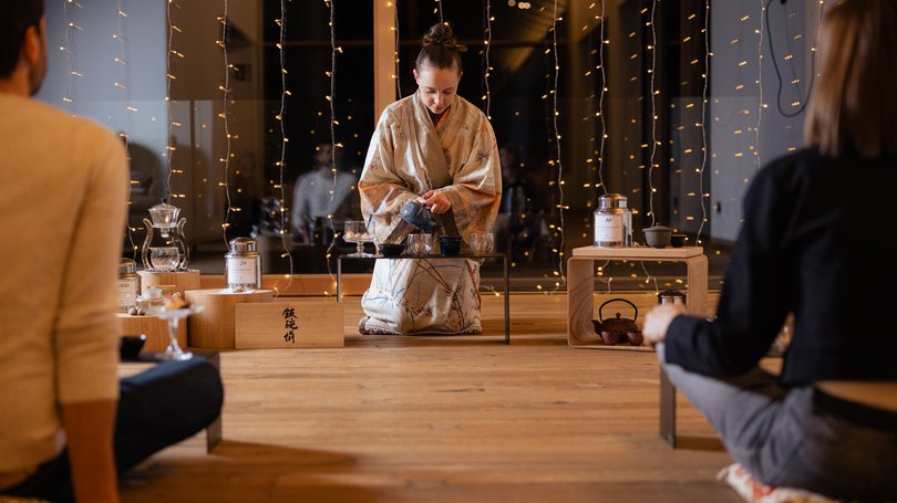 Escursioni da Moena e molto altro: il nostro programma Donna in kimono che prepara una cerimonia del tè giapponese davanti a due persone sedute.