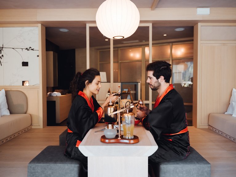 Val di Fassa in estate o inverno? Il relax non ha tempo! Coppia in kimono nero e rosso seduta a un tavolo in una stanza minimalista
