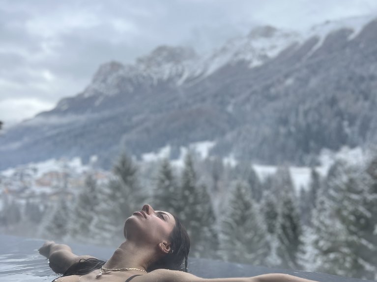 Val di Fassa in estate o inverno? Il relax non ha tempo! Donna rilassata in piscina a sfioro con vista su montagne innevate