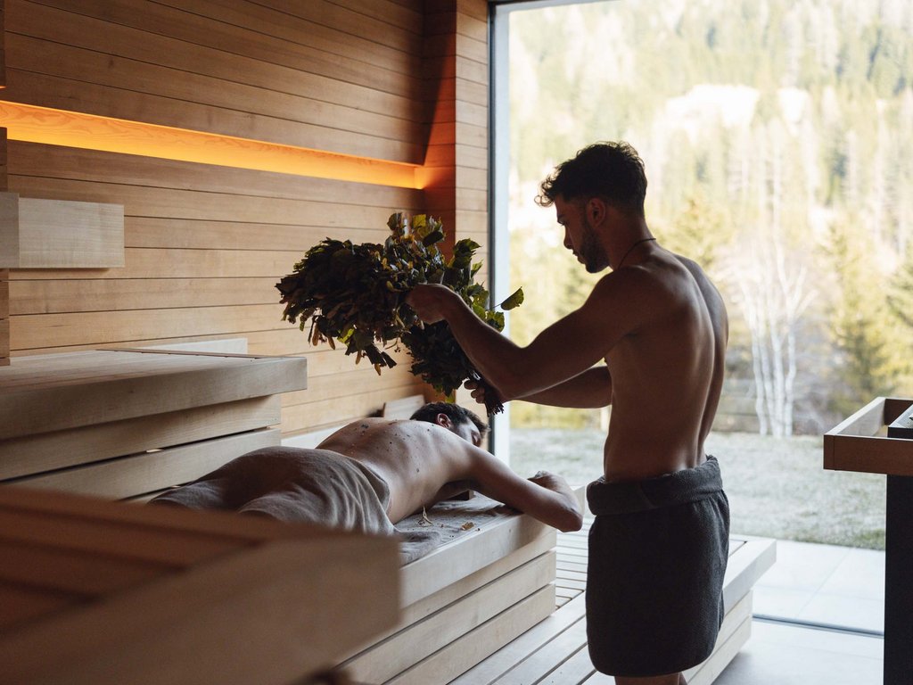 Al Latemar, dopo le passeggiate: relax nella Spa Oniro Due uomini nella sauna, uno disteso mentre l'altro agita rami verdi sopra di lui
