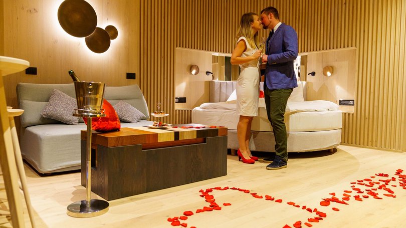 La pista ciclabile in Val di Fassa? C’è molto di più! Coppia romantica in suite d'albergo con petali di rose a forma di cuore sul pavimento.