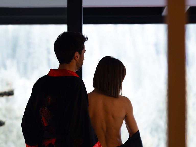 Val di Fassa in estate o inverno? Il relax non ha tempo! Coppia in accappatoi neri e rossi guardando fuori dalla finestra