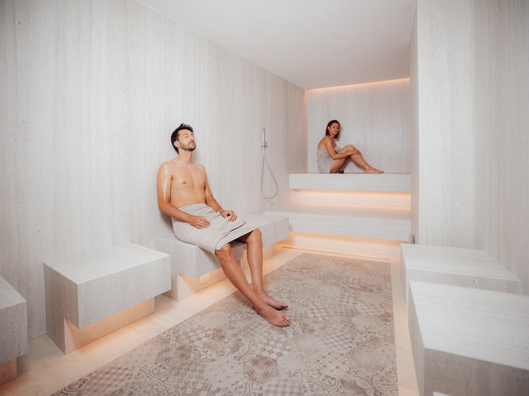 Il rituale dell’hammam al Latemar Hotel Uomo e donna rilassati in sauna moderna con pareti e pavimento chiari
