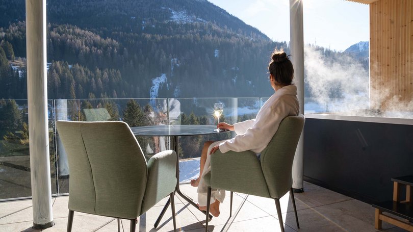 La pista ciclabile in Val di Fassa? C’è molto di più! Donna in accappatoio che si rilassa su terrazza con vista montagna e vasca idromassaggio