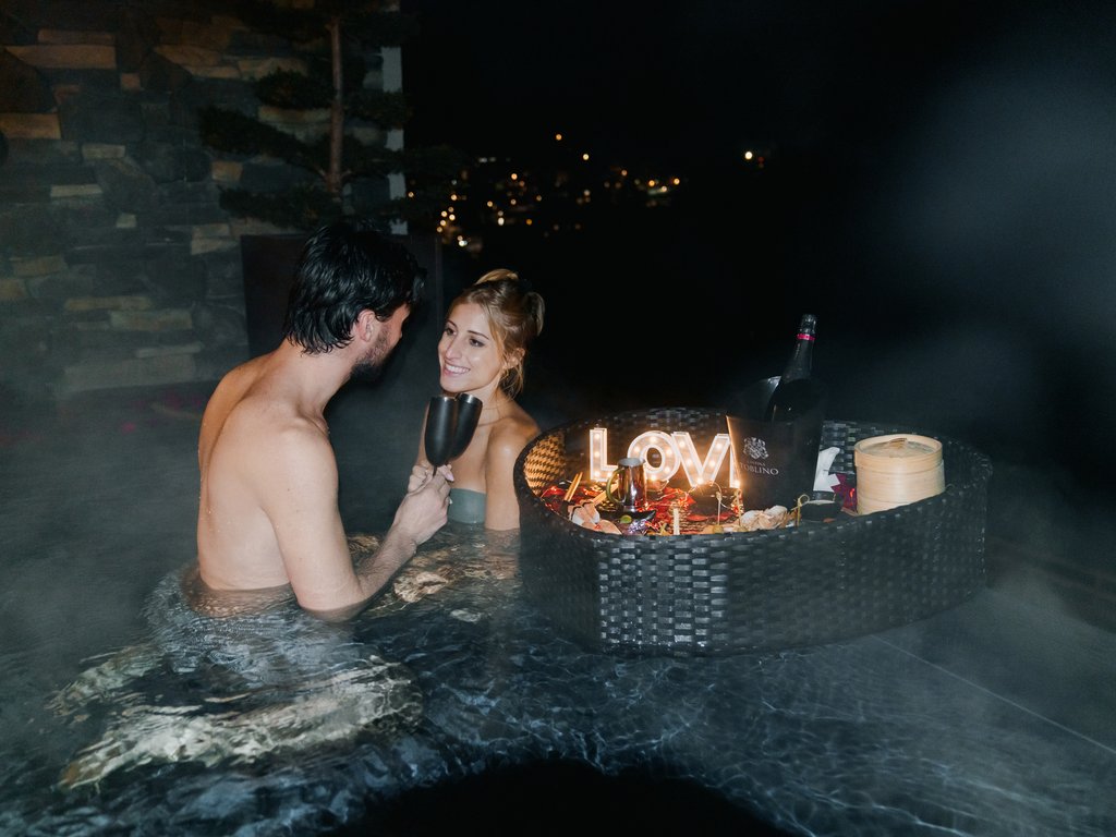 Val di Fassa in estate o inverno? Il relax non ha tempo! Coppia in vasca idromassaggio notturna con bicchieri e cena romantica galleggiante