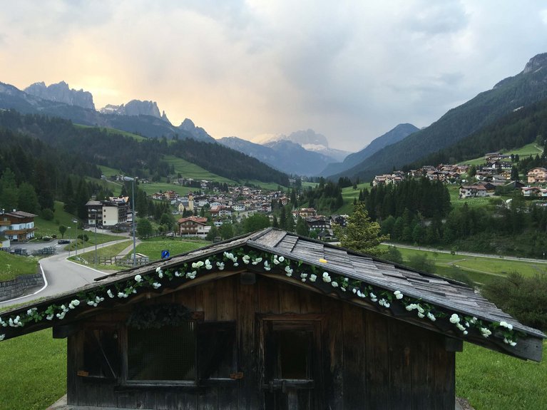 La pista ciclabile in Val di Fassa? C’è molto di più! Paesaggio montano con casetta decorata e villaggio in valle, cielo nuvoloso al tramonto