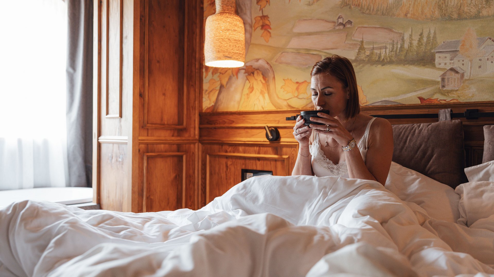 Hotel a 3 stelle superior a Soraga in Val di Fassa: camere & suites Donna seduta a letto accogliente che beve da una tazza nera in stanza rustica