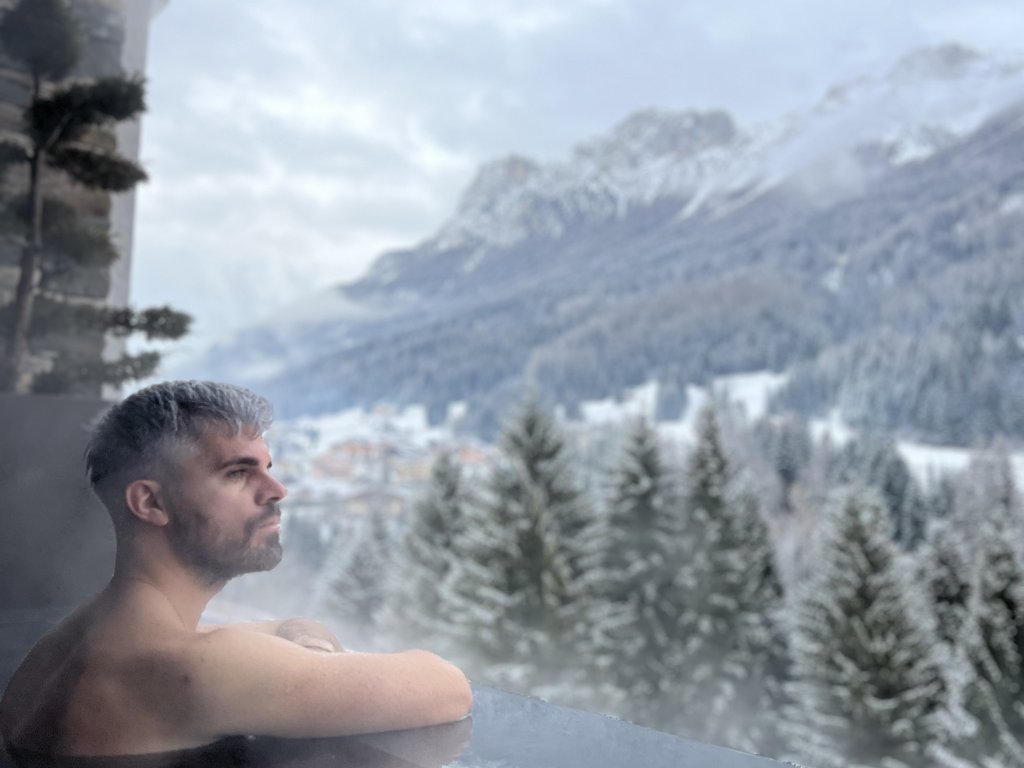 Val di Fassa in estate o inverno? Il relax non ha tempo! Uomo rilassato in vasca idromassaggio con vista sulle montagne innevate