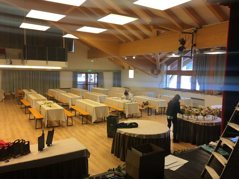 Dalla webcam il Latemar è bello, ma dal vivo è un sogno! Sala con tavoli e panche preparata per evento, due persone sistemano decorazioni