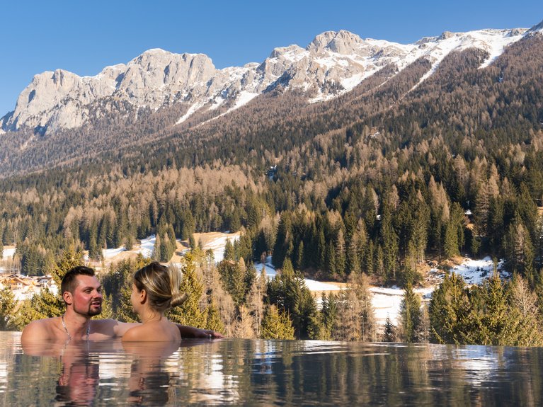 Val di Fassa in estate o inverno? Il relax non ha tempo! Coppia in piscina con vista sulle montagne innevate e foresta