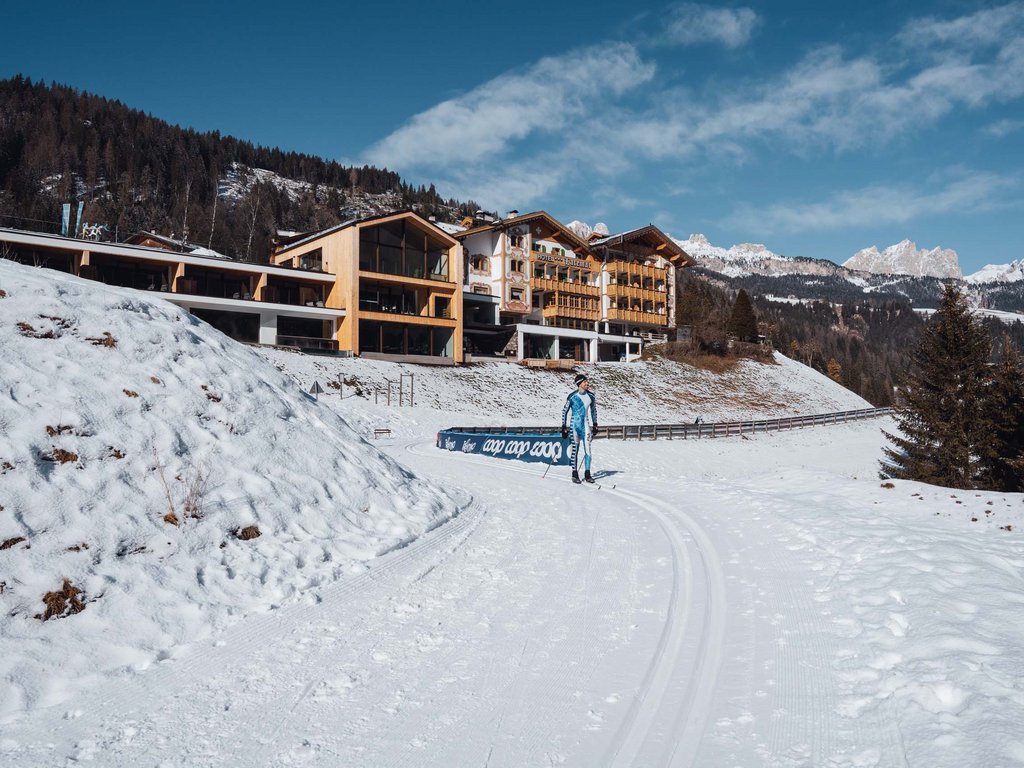 Passeggiate invernali in Val di Fassa: Hotel Latemar Sciatore su pista da sci di fondo davanti a hotel in montagna innevata