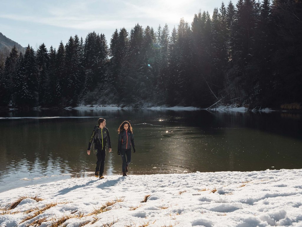 Passeggiate invernali in Val di Fassa: Hotel Latemar Due persone camminano sulla neve vicino a un lago circondato da alberi innevati
