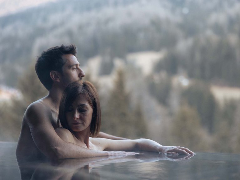 Val di Fassa in estate o inverno? Il relax non ha tempo! Coppia che si abbraccia in una piscina con vista sulle montagne