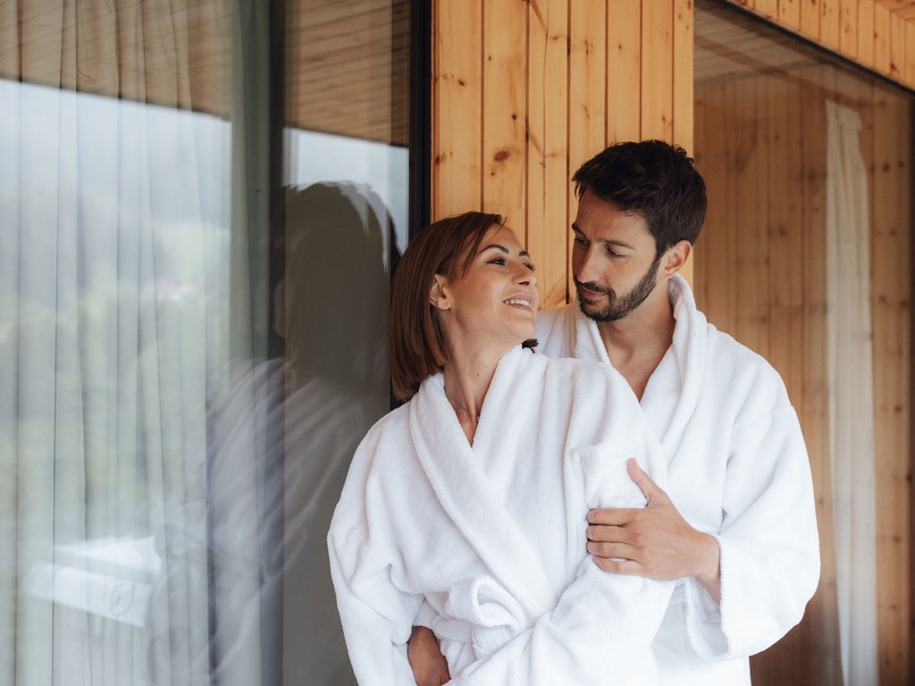 Moena, alberghi da sogno, panorama? C’è l’Hotel Latemar! Coppia in accappatoi bianchi abbracciata vicino a una finestra in casa in legno