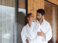 Dalla webcam il Latemar è bello, ma dal vivo è un sogno! Coppia in accappatoi bianchi abbracciata vicino a una finestra in casa in legno