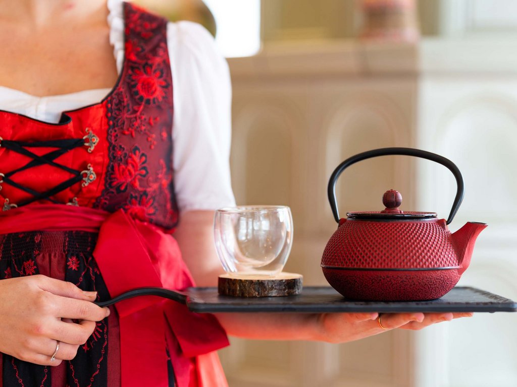 Val di Fassa e ciclabile: da ammirare dal cocktail & tea bar Donna in vestito tradizionale che porta un vassoio con teiera rossa e bicchiere