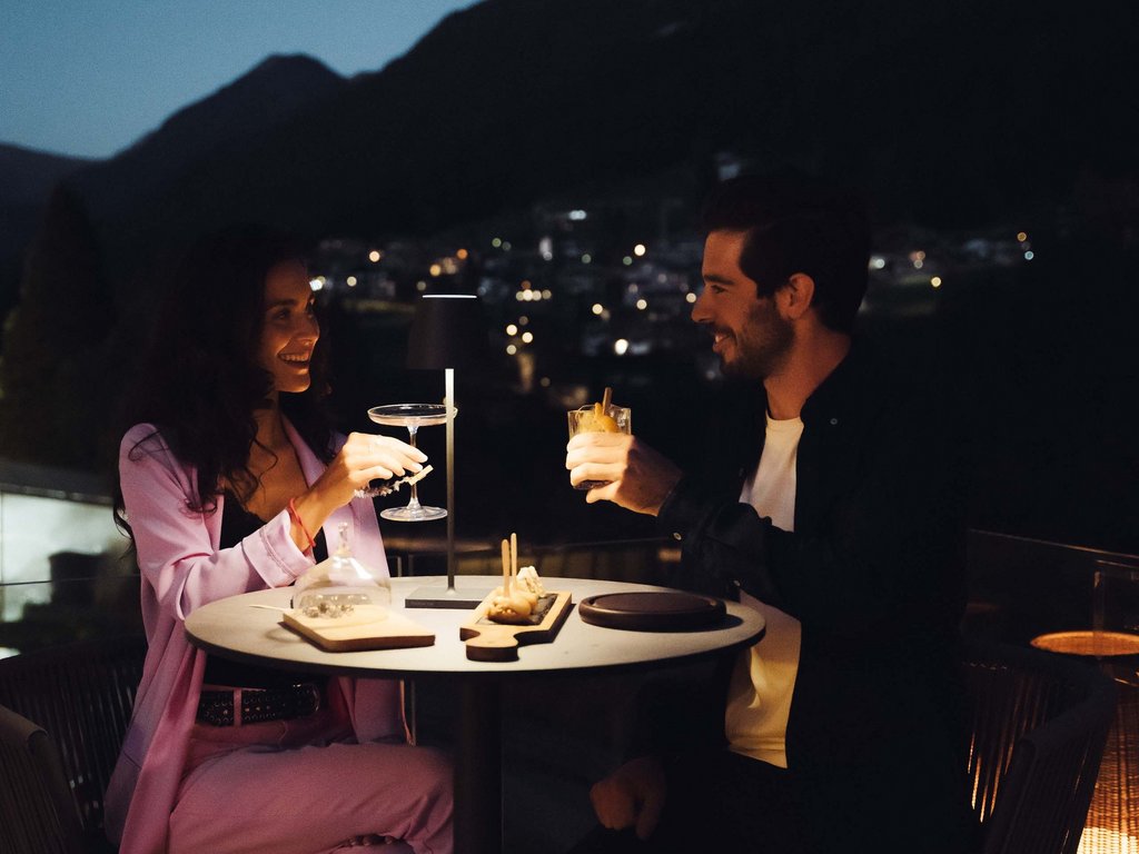 Hotel vicino a Moena a 4 stelle superior: relax con vista Coppia che brinda con cocktail a cena all'aperto di sera con luci soffuse.