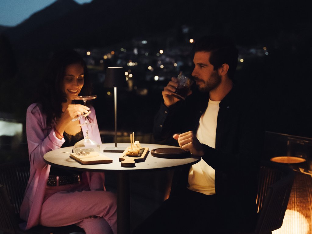 On the cycle track, from Moena to your romantic oasis Couple dining and drinking on a romantic terrace at night with mountains in the background